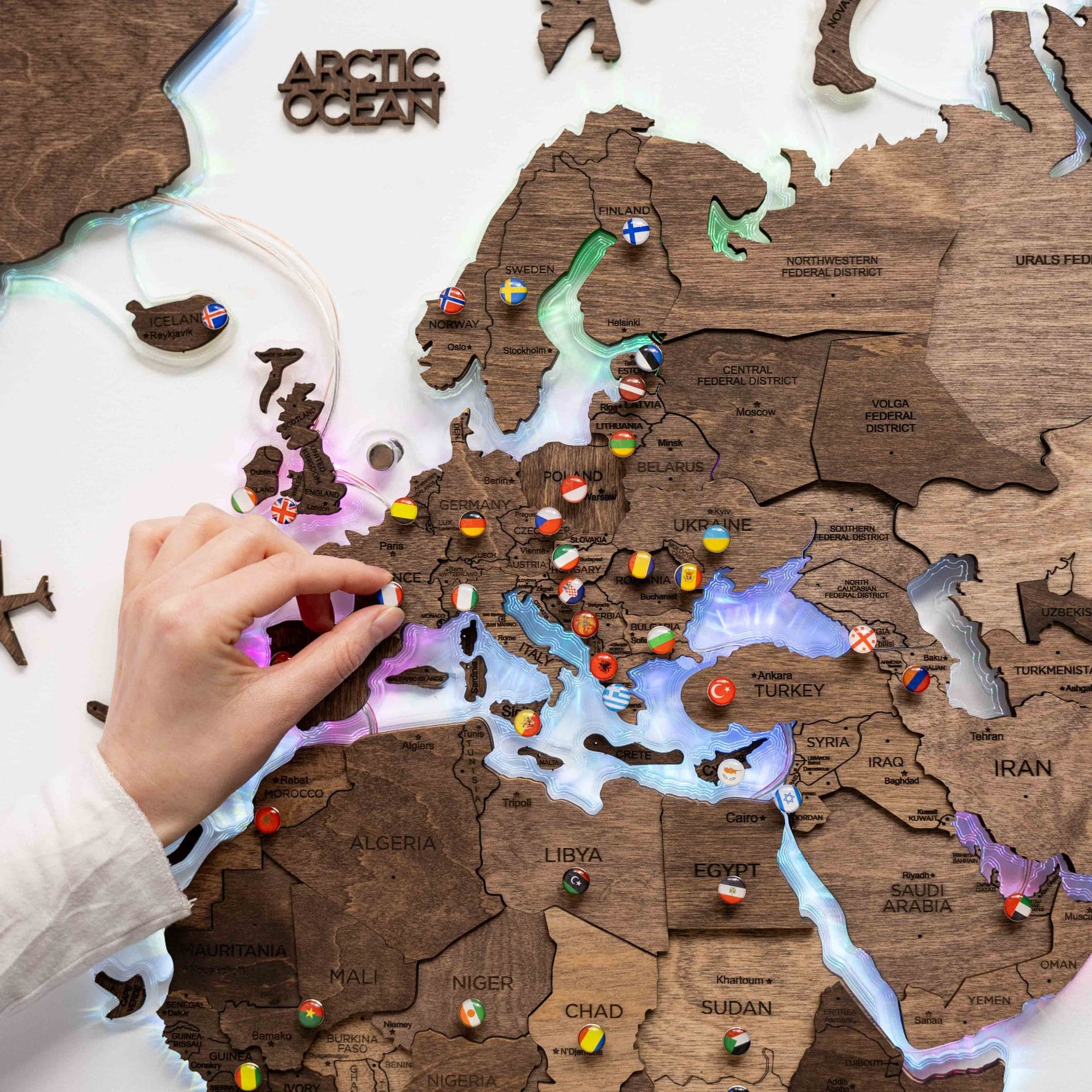 Wooden map of Europe with pins marking locations, hand pointing at a pin.